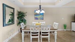 Uma área de refeições com mesa de madeira e cadeiras brancas, luminária pendente, um quadro na parede e um vaso com planta.