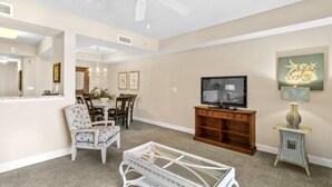 A living room with a ceiling fan, a television, a dining area with chairs and a table, and a decorative wall piece.