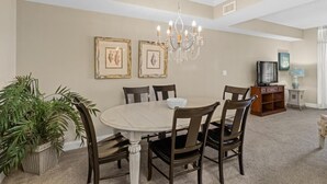 A dining area with a round table, six chairs, a chandelier, framed artwork on the wall, a television, and a side table with a lamp.