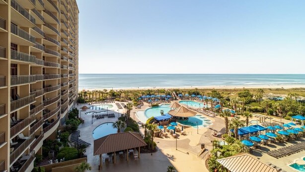 A hotel with multiple balconies, a pool area with various pools, and a beach view.
