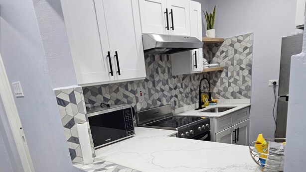 A modern kitchen with white cabinets, a marble countertop, and a built-in stove. There are two black stools and a small island with a sink.