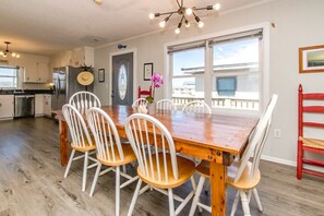 A dining area with a wooden table and white chairs, a kitchen in the background, and a view through large windows.