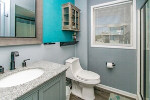 Une salle de bain avec un double vasque, des toilettes, un meuble, un miroir, une fenêtre avec des stores et un petit meuble au-dessus de la vasque.
