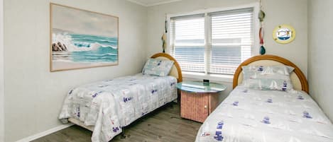 A bedroom with two beds, a painting of the ocean, a window with blinds, and a small round table.