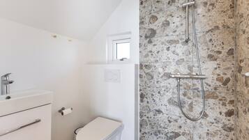 A bathroom with a white toilet, a sink with a faucet, a shower with a stone wall, and a window above the toilet.