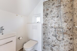 A bathroom with a white toilet, a sink with a faucet, a shower with a stone wall, and a window above the toilet. - Bilberghus - Beyond Bed & Breakfast (Hvide Sande)