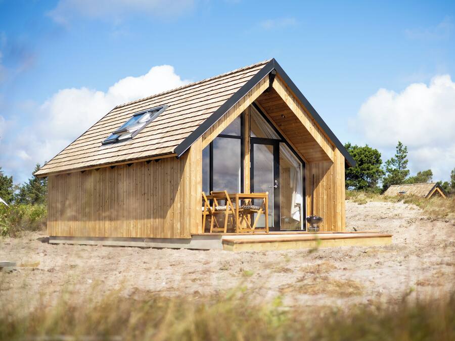 Een houten blokhut met schuin dak, grote ramen en een klein terras met stoelen.