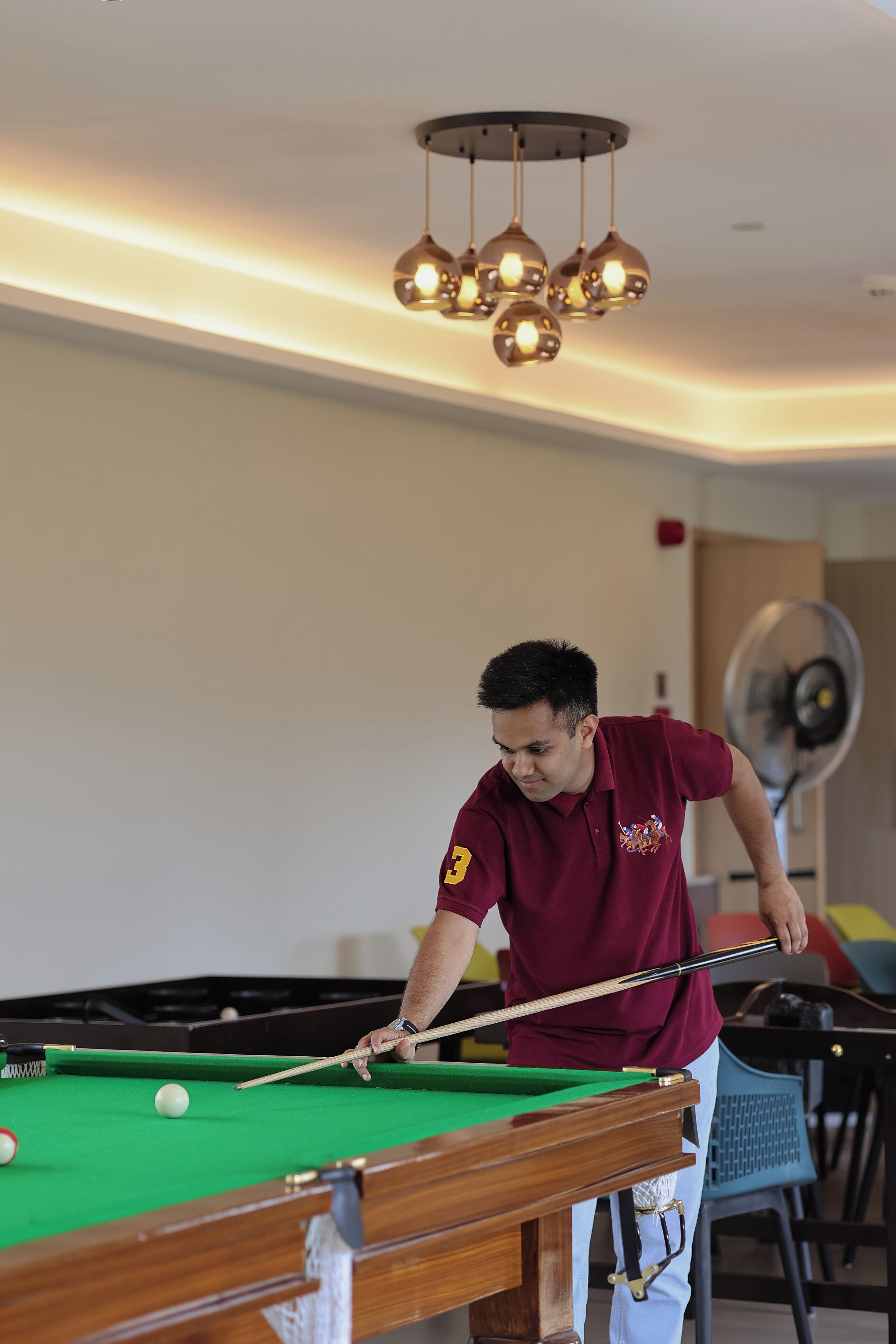 Eine Person spielt Billard in einem Raum mit einem grünen Billardtisch, einer Deckenleuchte und einem Ventilator im Hintergrund.