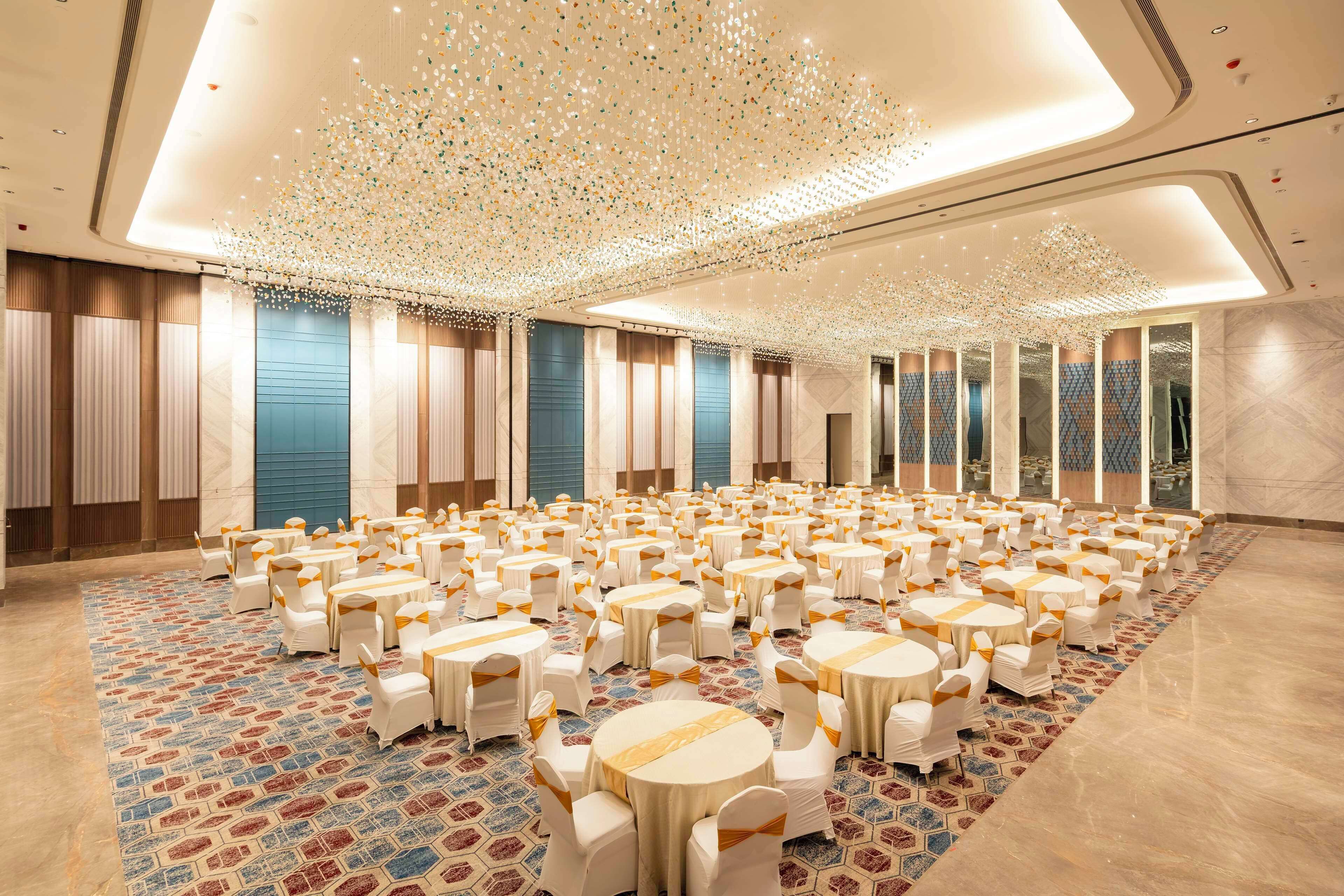A spacious banquet hall with round tables and white chairs, a patterned carpet, and a ceiling with hanging lights.