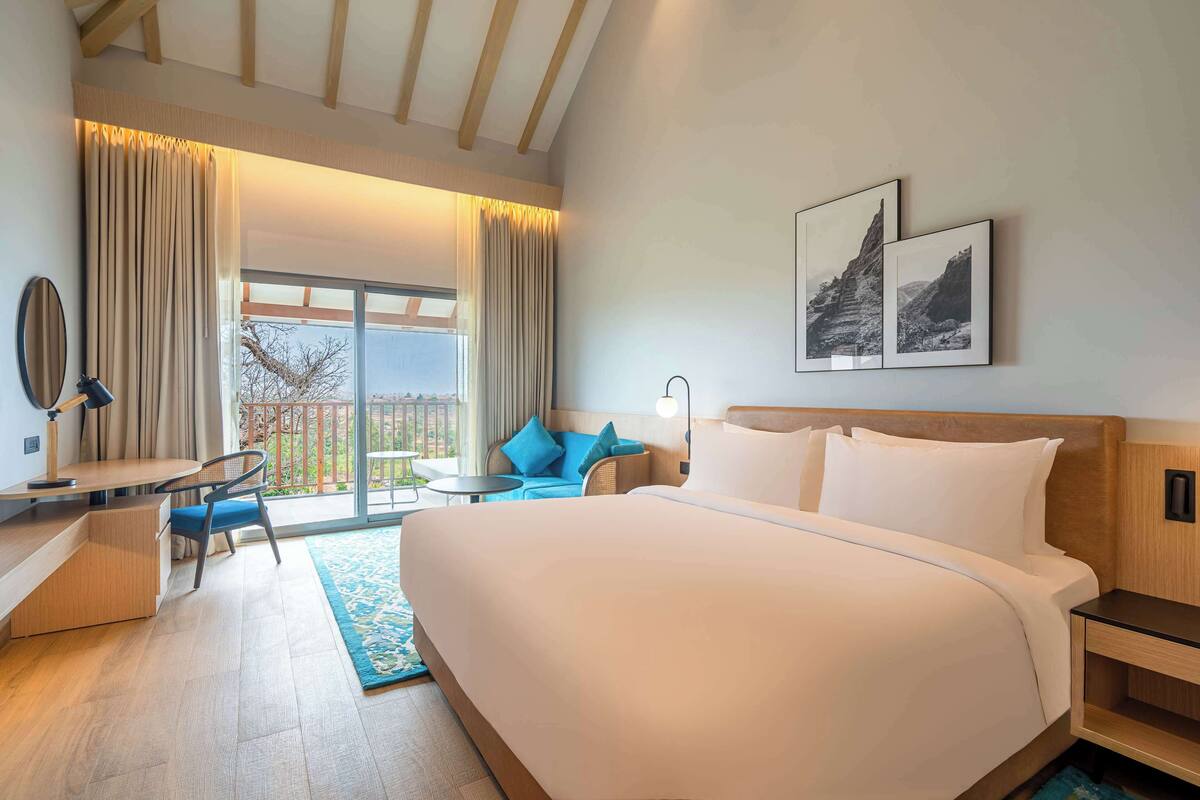 Quarto de hotel com uma cama grande, uma escrivaninha com cadeira, uma mesa pequena e uma varanda com vista.