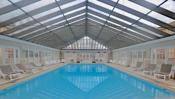 A large indoor swimming pool with a glass ceiling, surrounded by lounge chairs and a tiled floor.
