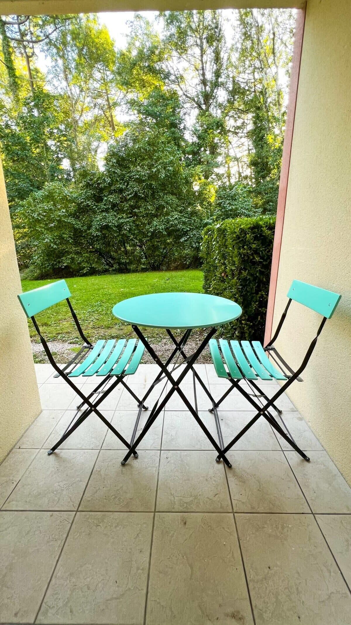 A patio area with a round table and two chairs, surrounded by a tiled floor and a view of a garden with trees and grass.