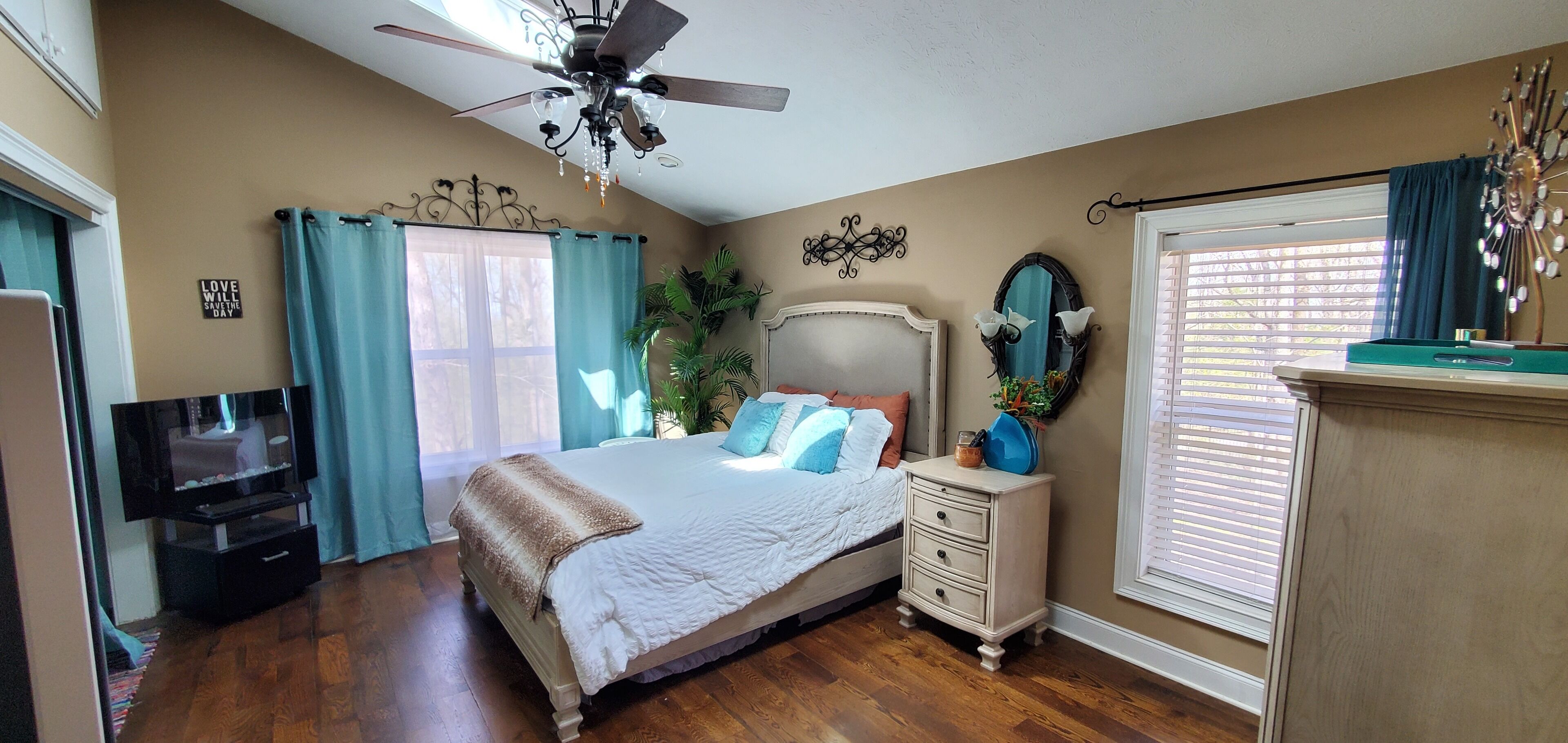 A bedroom with a bed, a nightstand, a television, a ceiling fan, a window with curtains, and a wall clock.