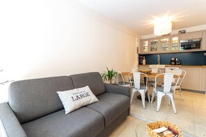 Un salon moderne avec un canapé gris, une table à manger avec des chaises blanches et un coin cuisine doté de meubles et d’appareils électroménagers.
