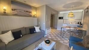 Una sala de estar moderna con un sofá gris, un área de comedor con una mesa pequeña y sillas, y una cocina al fondo.