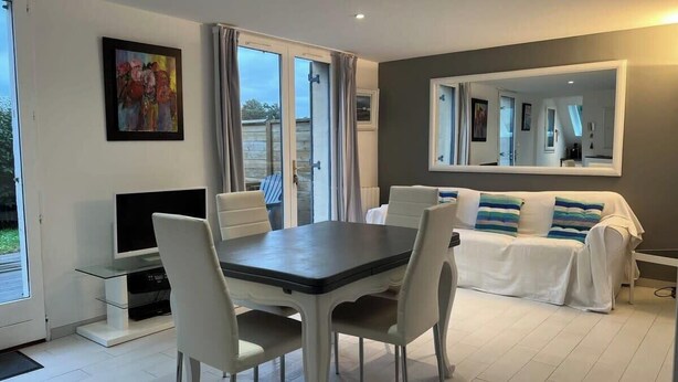 Un salon moderne comprenant une table à manger, des chaises blanches, un canapé avec des coussins à rayures, un téléviseur à écran plat et un grand miroir.