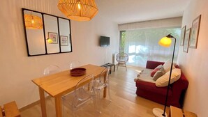 A modern living room with a wooden dining table, clear plastic chairs, a red sofa, and a large window.