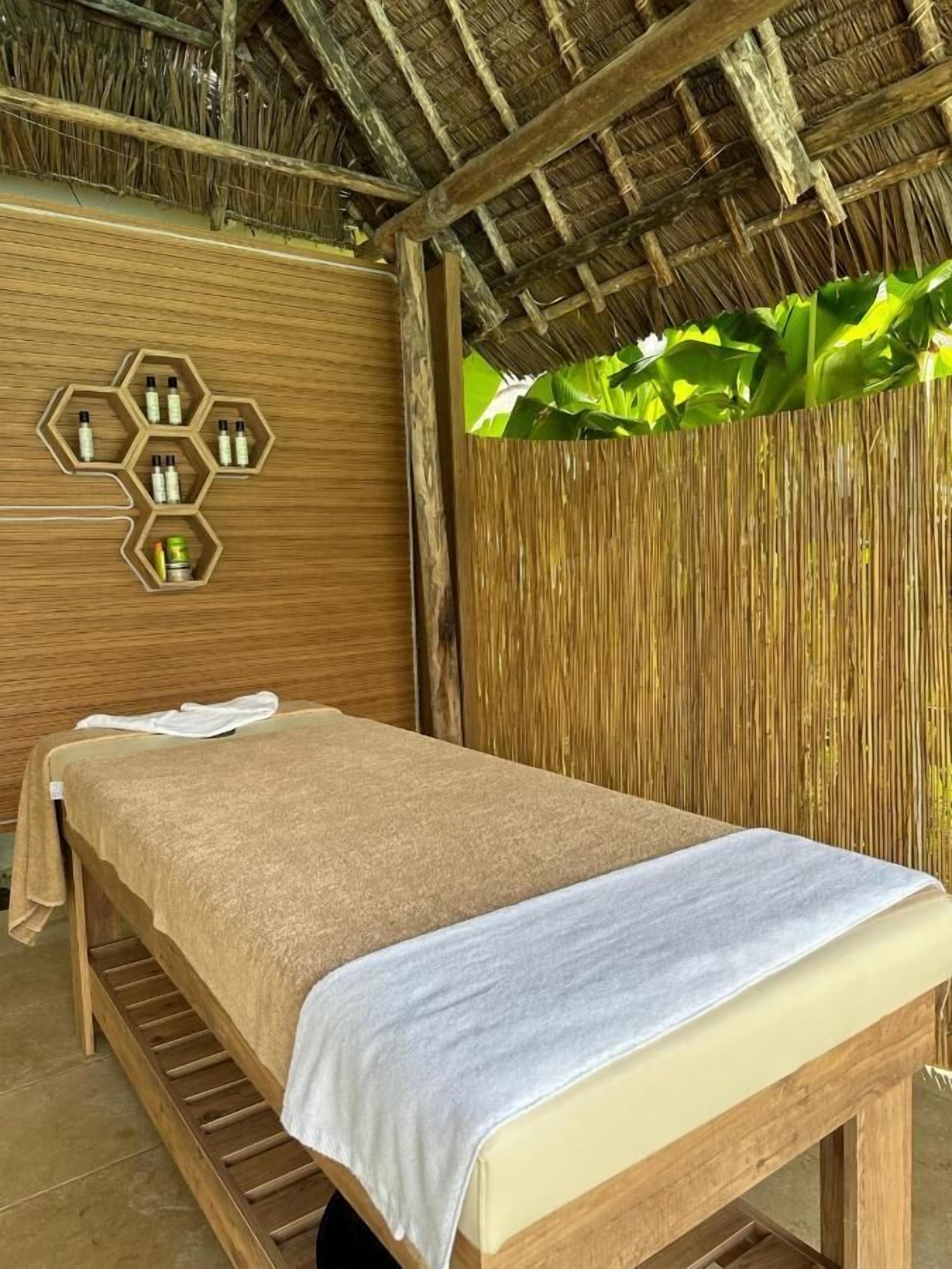 A wooden massage table with a beige towel and a white towel, set against a bamboo wall with a hexagonal shelf holding bottles.