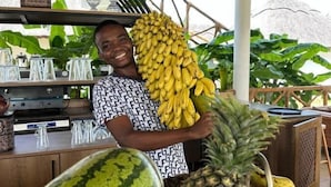 A person holding a large bunch of bananas and a pineapple, with a watermelon and a woven basket nearby. - Turkuaz Beach Hotel (Kiwengwa)