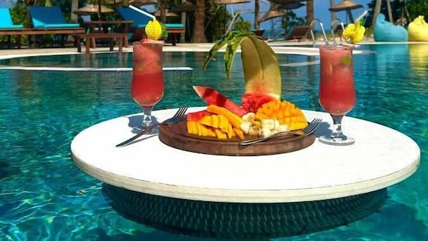 A poolside setting with a table featuring fruit and drinks, surrounded by palm trees and lounge chairs. - Turkuaz Beach Hotel (Kiwengwa)