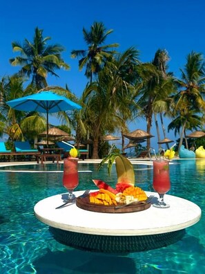 A poolside setting with a table featuring fruit and drinks, surrounded by palm trees and lounge chairs. - Turkuaz Beach Hotel (Kiwengwa)