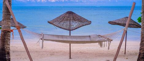 A hammock between two palm trees on a sandy beach with a clear view of the ocean.