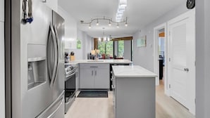 A modern kitchen with a stainless steel refrigerator, built-in oven, and a large island with a sink. There is a ceiling light fixture and a clock on the wall. - Award-Winning Resort Amenities | Shipwatch 223D (Isle of Palms)