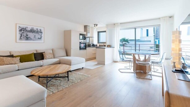 Ein modernes Wohnzimmer mit einer Couch, einem Couchtisch und einem Essbereich mit Stühlen. Im Hintergrund ist eine Küche zu sehen.