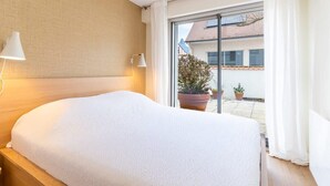 Ein Hotelzimmer mit einem großen Bett, zwei Nachttischlampen, einer Schiebetür zu einem Balkon mit Topfpflanzen und Blick auf ein Gebäude.