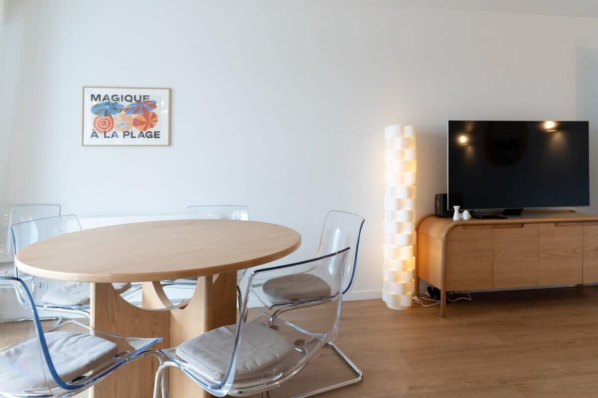 A modern living room with a round wooden table, clear glass chairs, a flat-screen TV on a wooden cabinet, and a wall-mounted light fixture.