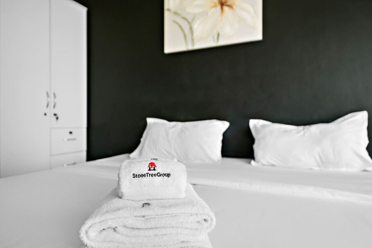 Un lit bien fait, avec du linge de lit et des oreillers blancs, une serviette blanche pliée ornée d’un logo, et une armoire blanche dans une pièce dont un mur est peint dans une couleur foncée et qui est décorée d’un tableau représentant des motifs floraux.