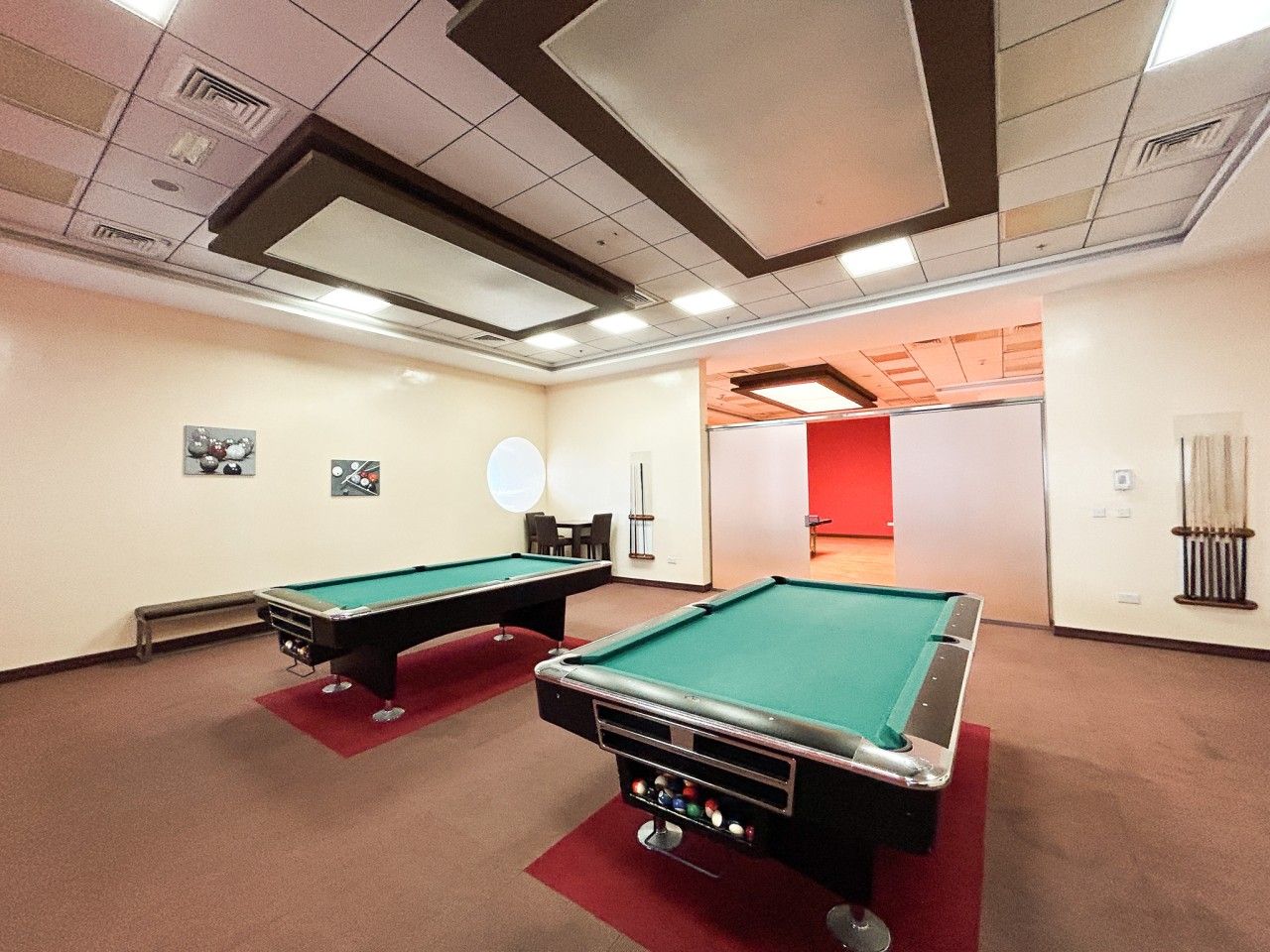 Une pièce avec deux tables de billard, une moquette rouge et un mur blanc orné d’un luminaire circulaire.