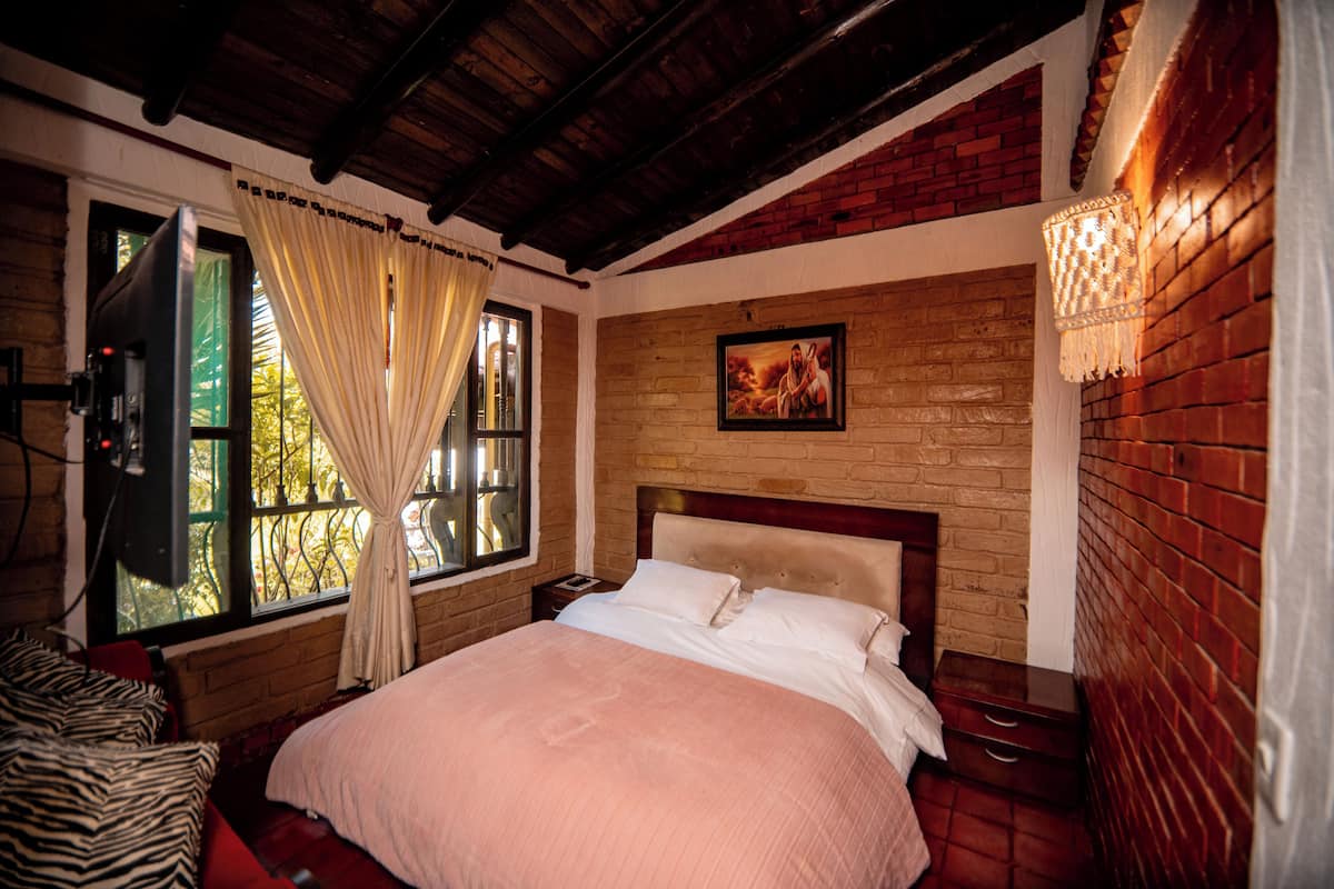 A bedroom with a brick wall, a wooden ceiling, a bed with white bedding, a nightstand, a lamp, and a window with curtains.