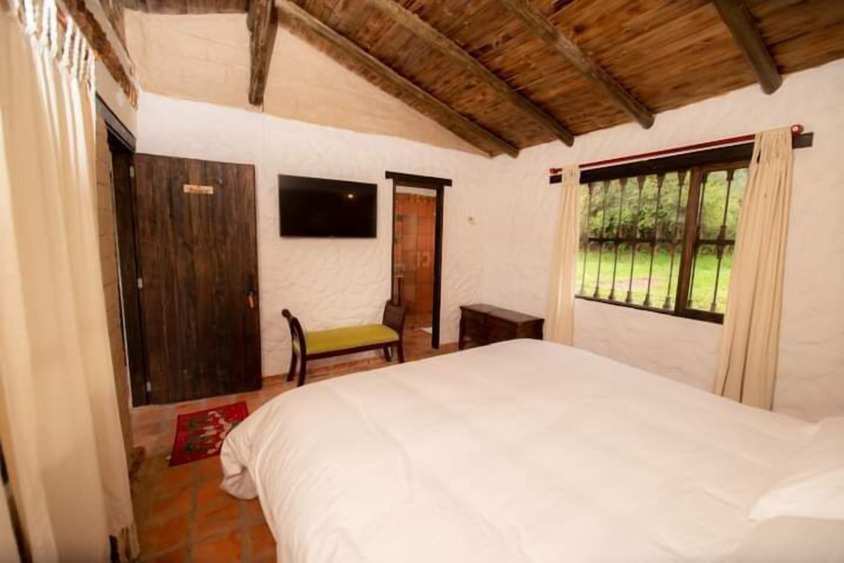 A bedroom with a bed, a wooden bench, a small table, a door, and a window with curtains.