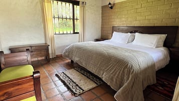 Un dormitorio con cama, cabecero de madera, mesita de noche, un banco, ventana con cortinas y pared de ladrillo.