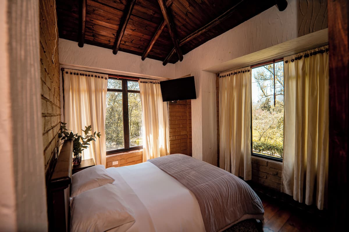 A bedroom with a wooden ceiling, a bed with white linens, a flat-screen TV mounted on the wall, and windows with curtains.
