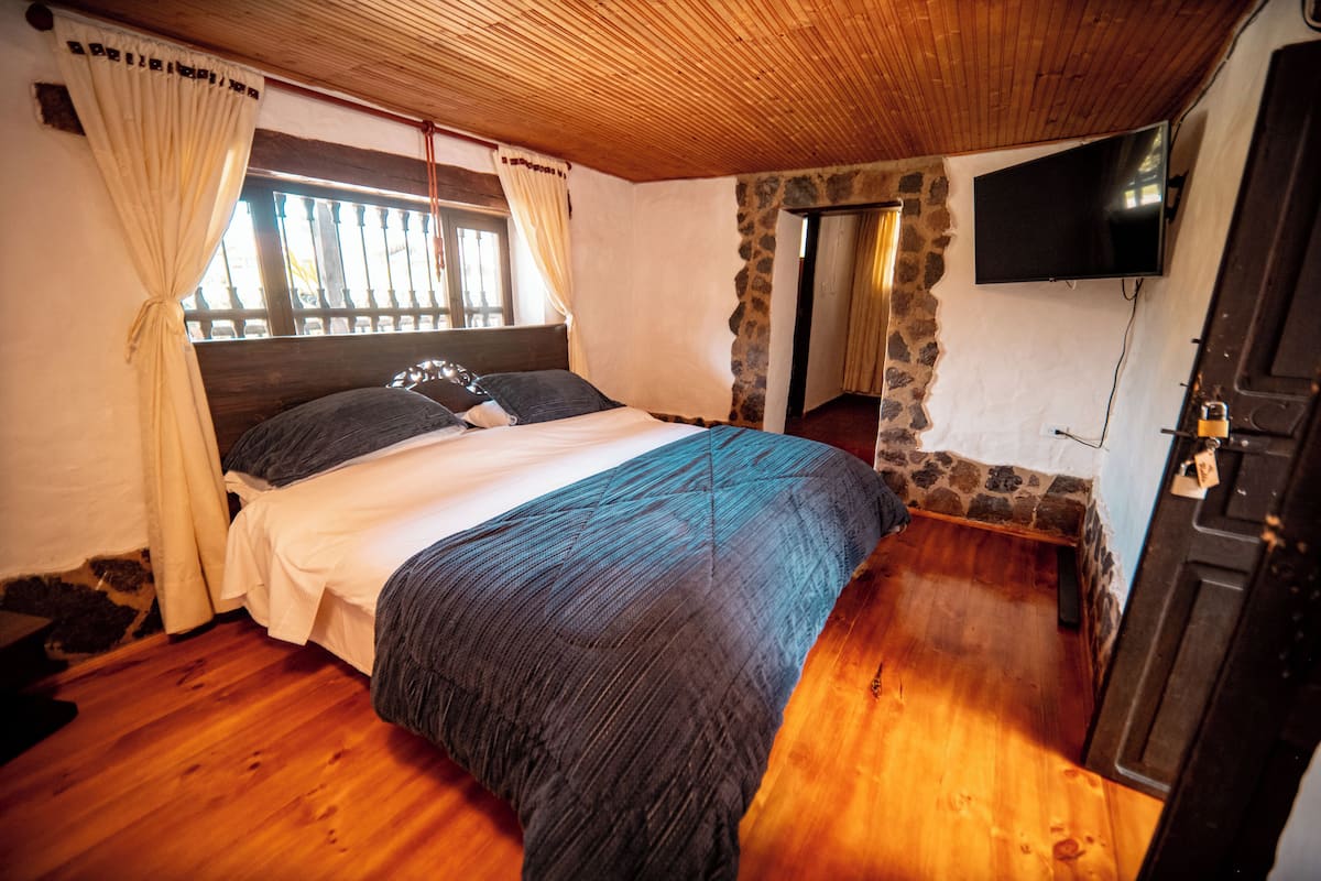 A bedroom with a wooden ceiling, a stone wall, a bed with a blue blanket, a TV mounted on the wall, and a wooden door.
