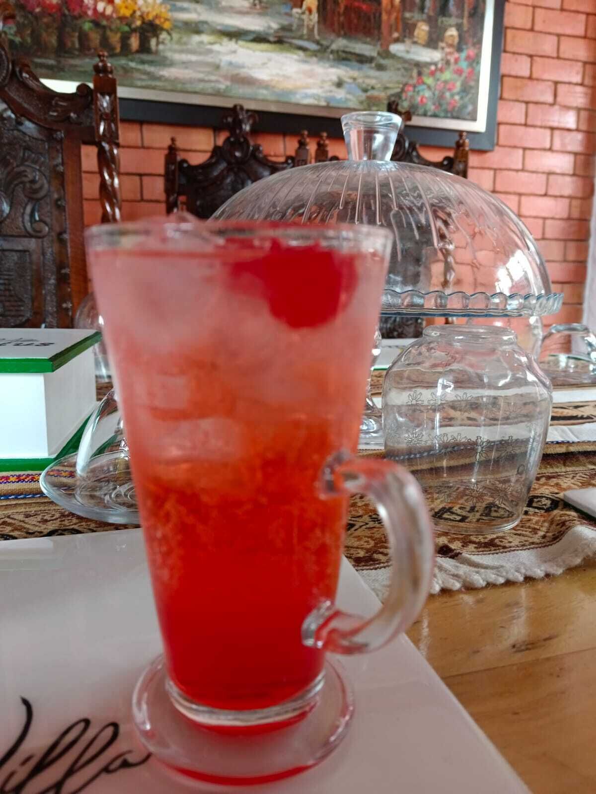 A clear glass pitcher with a handle, filled with a red beverage, placed on a table with a patterned tablecloth.