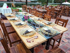 Una larga mesa de comedor dispuesta con sillas de madera, cristalería y posavasos tejidos.