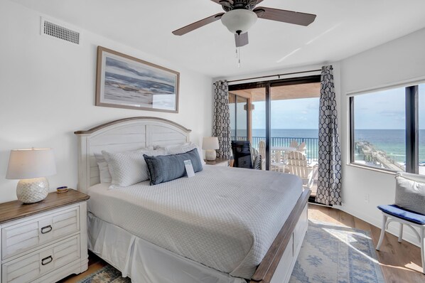 A bedroom with a bed, a nightstand, a lamp, a ceiling fan, a window with a view of the ocean, and a balcony with chairs.