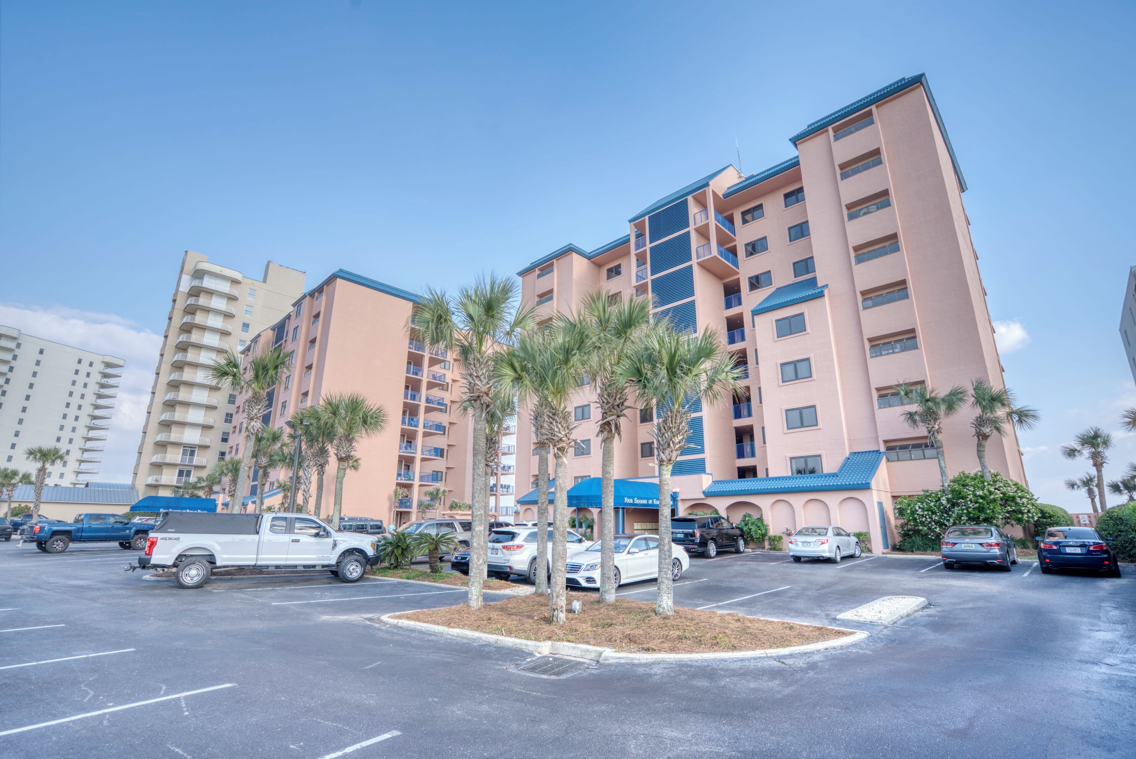A multi-story hotel with a parking lot, palm trees, and a clear blue sky.