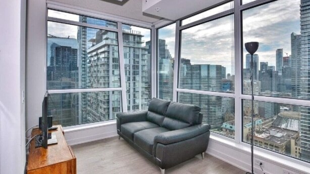A modern living room with a grey sofa, large windows offering a city view, and a wooden side table. - Studio 2BR Apt with GYM, Close to CN Tower (Toronto)