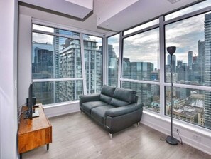A modern living room with a grey sofa, large windows offering a city view, and a wooden side table. - Studio 2BR Apt with GYM, Close to CN Tower (Toronto)