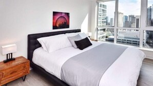 A modern bedroom with a large bed, a nightstand, a city view, and a framed picture on the wall. - Studio 2BR Apt with GYM, Close to CN Tower (Toronto)