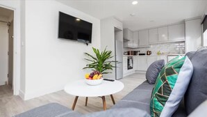 Ein modernes Wohnzimmer mit grauem Sofa, einem runden weißen Couchtisch mit Obstschale, einem Topfpflanzen und einem flachbildfernseher an der Wand.