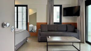 Ein modernes Hotelzimmer mit Bett, Sofa, Couchtisch, Fernseher und einem großen Fenster mit Vorhängen.