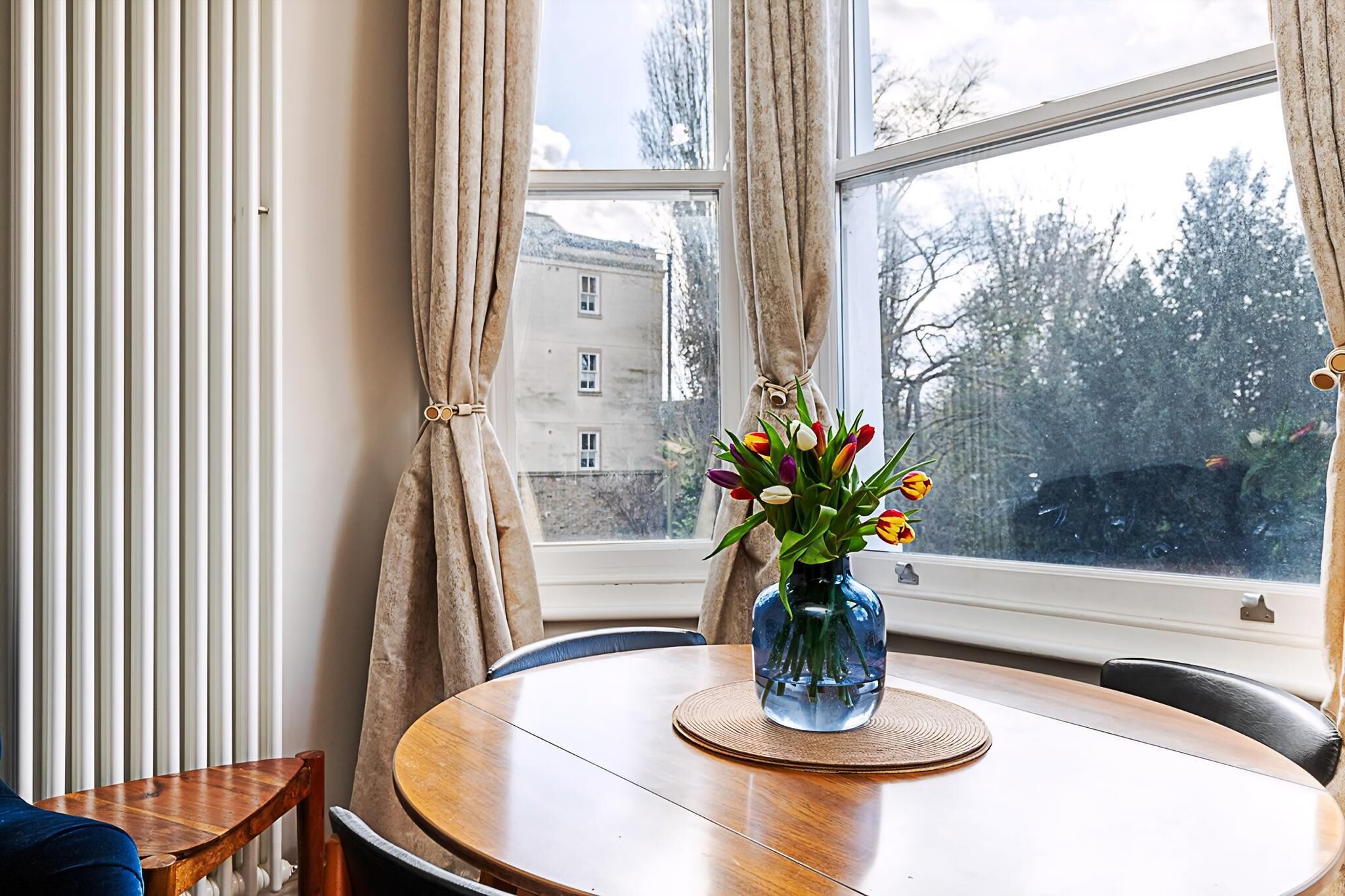 A round wooden table with a blue vase containing tulips, positioned by a window with curtains.