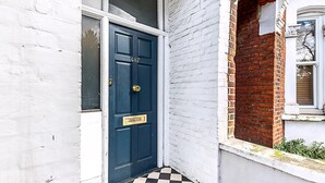 A white brick building with a dark blue door, a checkered floor, and a small window with a brick wall. - Lovely Bishops Park Flat (London)