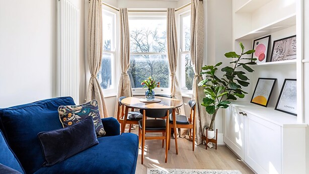 A cozy corner with a blue sofa, a dining table with chairs, a potted plant, and a bay window with curtains. - Lovely Bishops Park Flat (London)