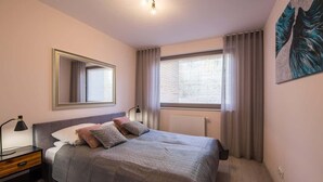 Ein Schlafzimmer mit Bett, Nachttisch, Lampe, einem Fenster mit Vorhängen, einem Heizkörper und einem an der Wand befestigten Spiegel.